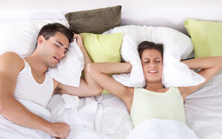 Couple in bed; man snoring and woman can not sleep; covering ears with pillow for snore noise.
